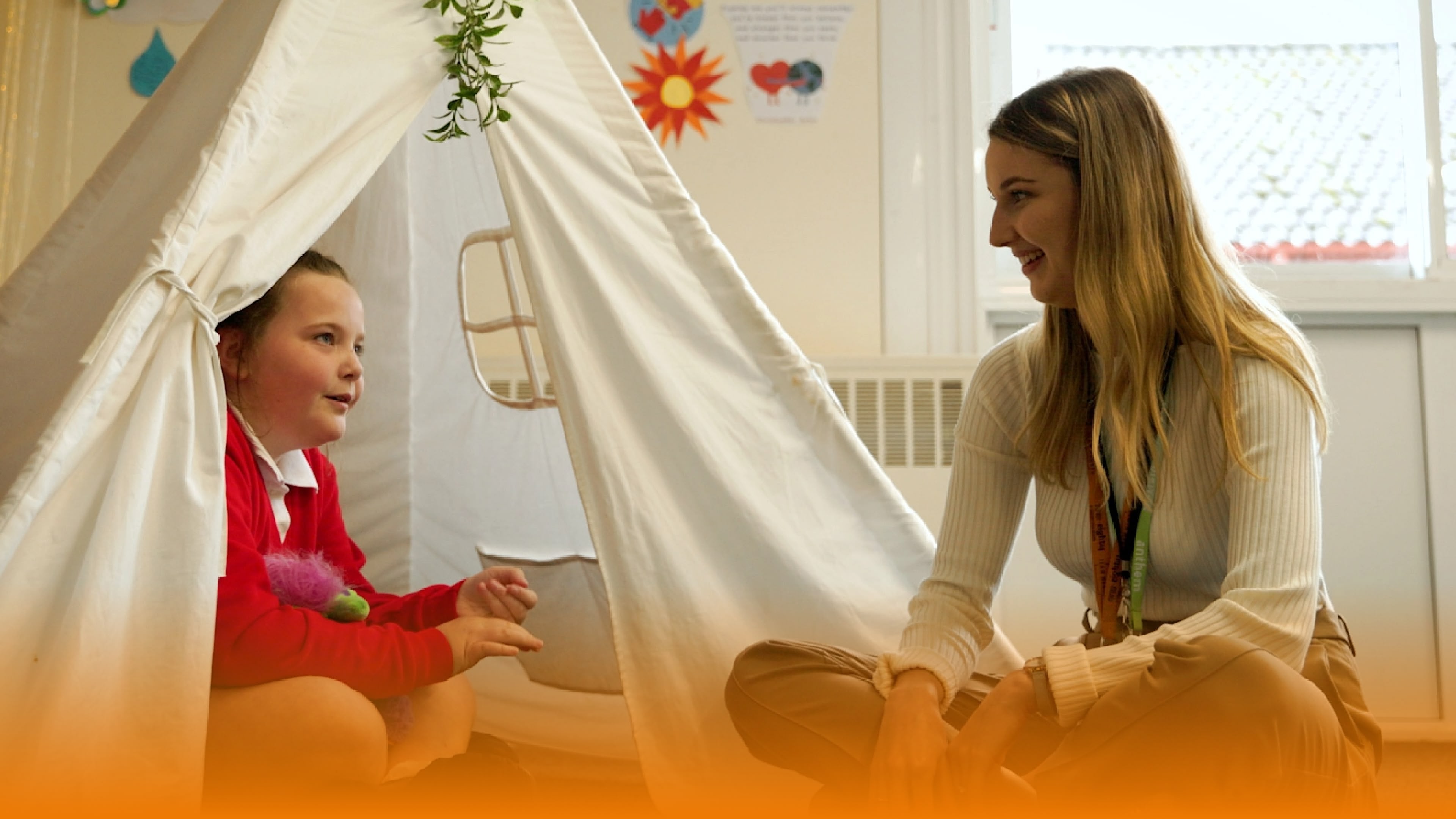 A young girl in a red top sits inside a white teepee while a woman sits nearby in a bright playroom.