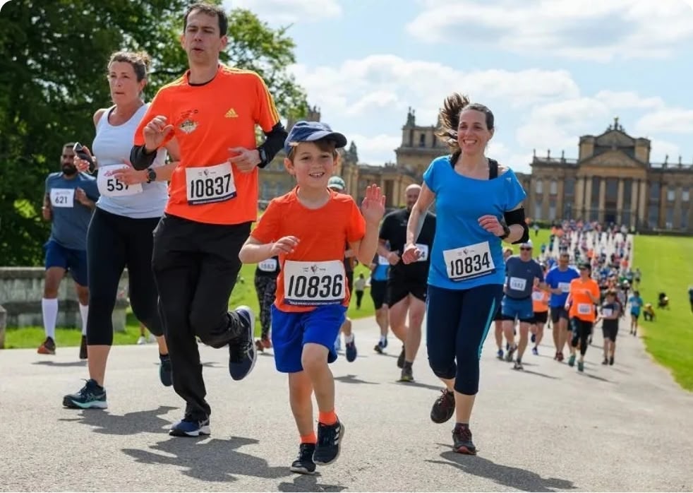 Group of runners participating in a sunny park race, including a young boy in an orange shirt with a bib and a woman in blue nearby.