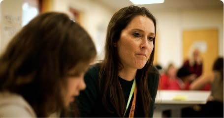 A woman with dark hair sits at a table in a classroom, looking toward the camera with a thoughtful expression while blurred figures are seen in the background.