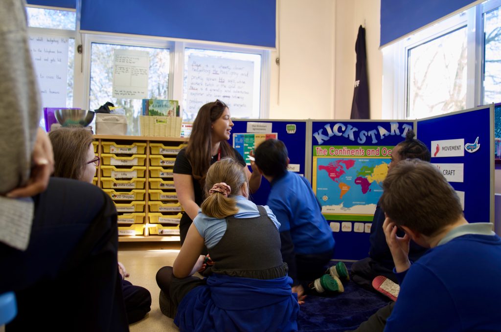 Children sit on the floor in a classroom around a presenter beside a blue display board featuring a world map.