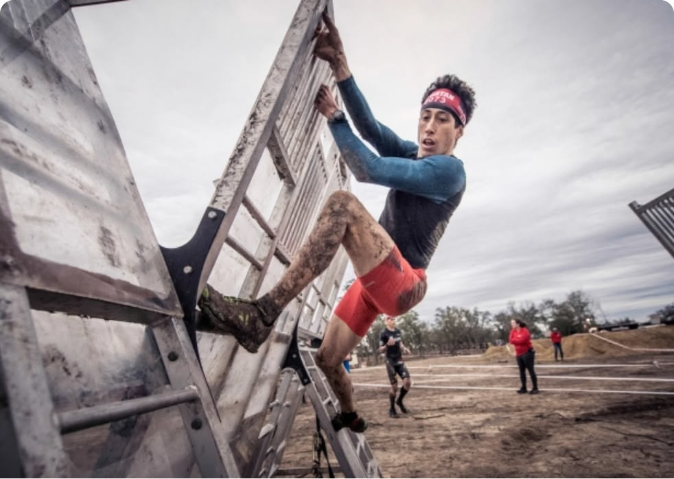 A person in a blue long-sleeve shirt and red shorts climbs over a wooden obstacle wall at an outdoor obstacle race.